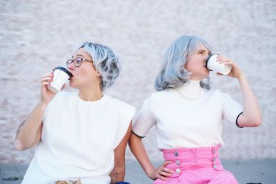 Szene aus dem Stück „Die Nashörner. Oder. Ein Duell für Leichtgläubige.“: Die Kandidatinnen Wisser und Stech sitzen nebeneinander auf einer Bank, schauen in entgegengesetzte Richtungen und trinken Coffee to go.